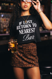 A woman at a bar, wearing sunglasses and an animal print skirt, holds a drink. She sports the Panache Apparel Collection’s “If Lost, Return To Nearest Bar Tee,” featuring a bold black graphic design. Shelves filled with bottles are blurred in the background.