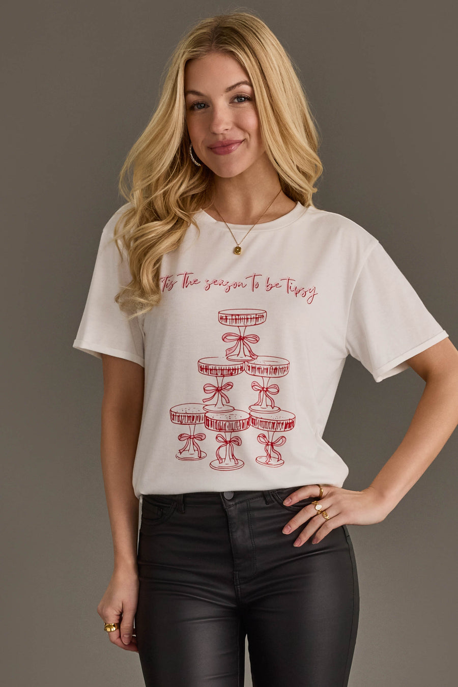 A woman wears a Tis The Season To Be Tipsy Tee, featuring a white shirt with a red line-art illustration of a champagne coupe tower and the text Tis the season to be tipsy.