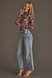 A woman poses in light-wash wide-leg jeans, nude heels, and the Olive & Pink Floral Mesh Top from Panache Apparel Collection against a plain gray background, smiling with one hand near her face.