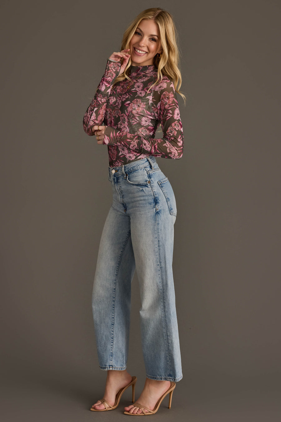 A woman poses in light-wash wide-leg jeans, nude heels, and the Olive & Pink Floral Mesh Top from Panache Apparel Collection against a plain gray background, smiling with one hand near her face.