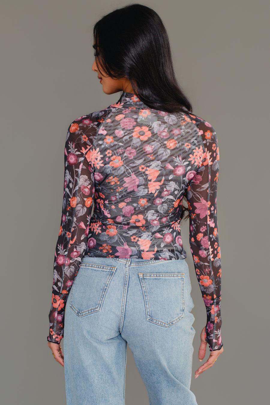 A woman with long dark hair is photographed from the back against a plain gray background, wearing the Panache Apparel Collection Black, Orange & Pink Floral Mesh Top paired with light blue jeans.