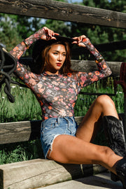 A woman wearing the Panache Apparel Collection Black, Orange & Pink Floral Mesh Top with long sleeves and a mock neckline, paired with denim shorts and black cowboy boots, sits on the ground by a wooden fence holding a hat, surrounded by green grass and trees.