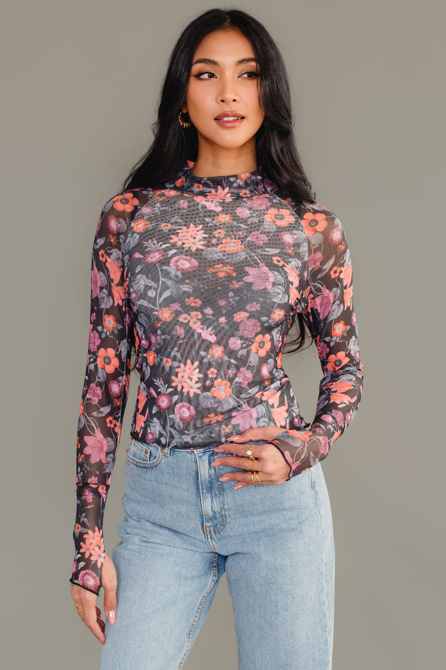 A woman models the Panache Apparel Collection Black, Orange & Pink Floral Mesh Top paired with blue jeans, standing against a plain gray background, looking slightly to her left with one hand on her hip.