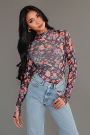 A woman with long black hair wears the Black, Orange & Pink Floral Mesh Top from Panache Apparel Collection and light blue jeans, standing against a plain gray background and smiling slightly.