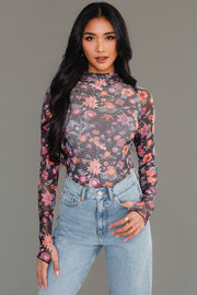 A woman with long black hair, dressed in the Panache Apparel Collection’s Black, Orange & Pink Floral Mesh Top and high-waisted light blue jeans, stands against a plain gray background, looking confidently at the camera.