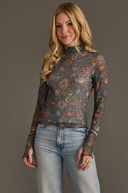 A woman with long blonde hair wears the Panache Apparel Collection’s Gray, Orange & Blue Floral Mesh Top paired with light blue jeans, standing against a plain neutral background and smiling softly.