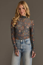 A woman with long blonde hair wears the Panache Apparel Collection’s Gray, Orange & Blue Floral Mesh Top paired with light blue high-waisted jeans, posing against a plain gray background and smiling softly at the camera.