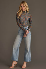A woman stands against a plain background, wearing the Panache Apparel Collection’s Gray, Orange & Blue Floral Mesh Top paired with light blue wide-leg jeans and nude high-heeled sandals. She has long blonde hair and is smiling slightly.