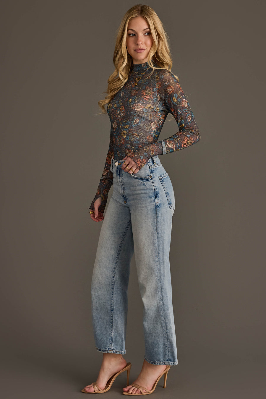 A woman poses against a plain gray background wearing the Panache Apparel Collection’s Gray, Orange & Blue Floral Mesh Top, a sheer, long-sleeve piece paired with light blue jeans and nude high-heeled sandals.
