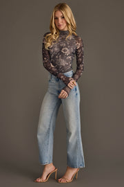 A woman stands against a plain background wearing the Panache Apparel Collection’s Gray & Cream Floral Mesh Top, paired with light blue high-waisted jeans and nude heeled sandals. She has long blonde hair and poses with one hand on her hip.