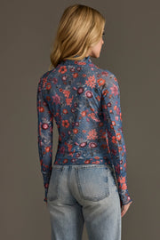 A person with long blonde hair, wearing the Panache Apparel Collection’s Blue & Orange Floral Mesh Top and light blue jeans, stands facing away from the camera against a plain gray background.