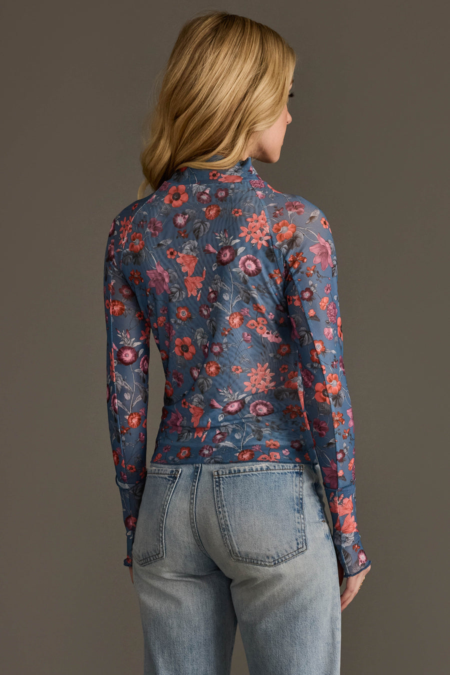 A person with long blonde hair, wearing the Panache Apparel Collection’s Blue & Orange Floral Mesh Top and light blue jeans, stands facing away from the camera against a plain gray background.