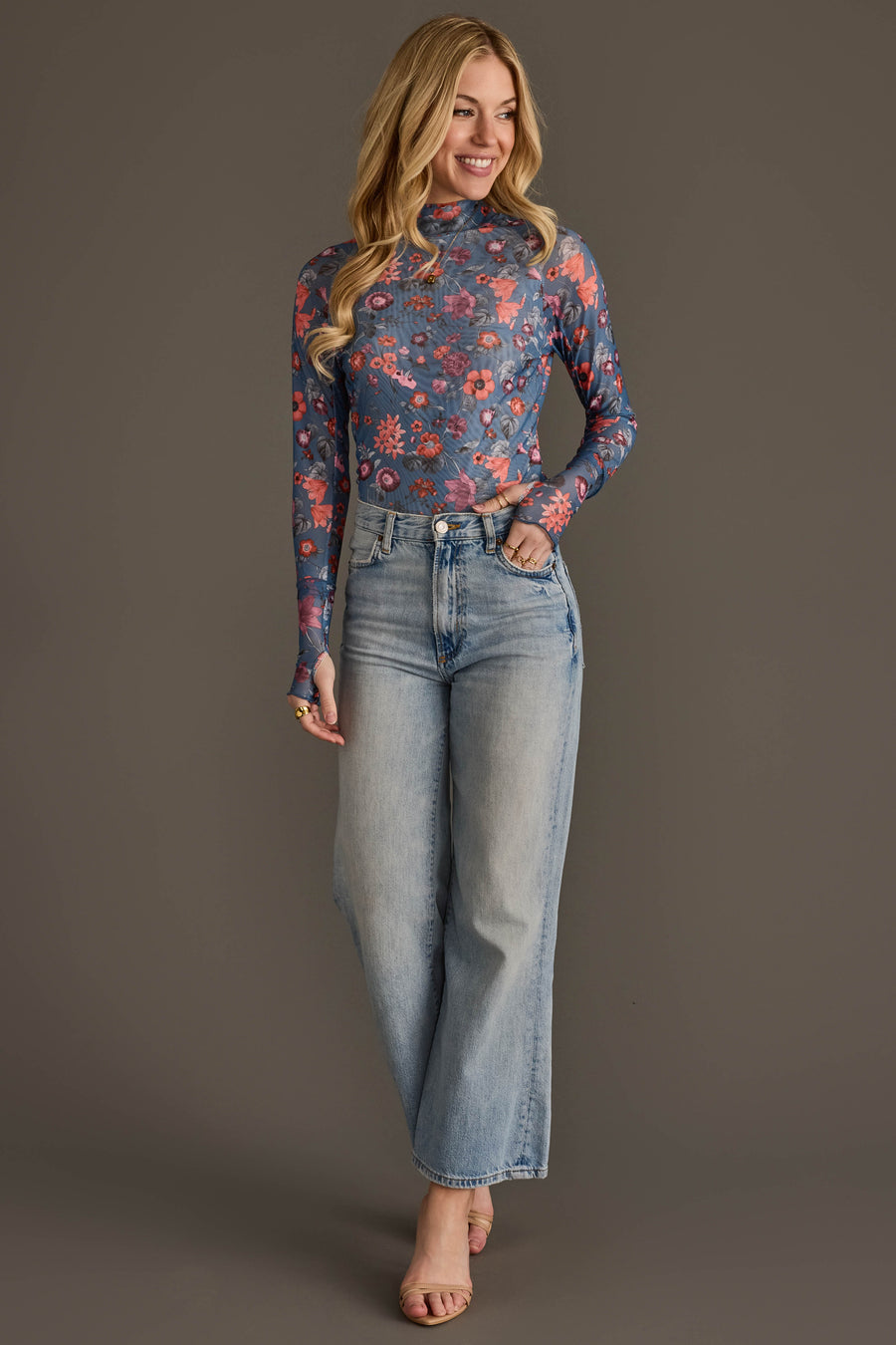 A woman with long blonde hair stands smiling with one hand in her pocket against a plain gray background. She wears the Panache Apparel Collection’s Blue & Orange Floral Mesh Top, styled with high-waisted light blue wide-leg jeans.