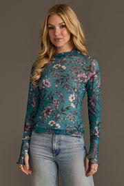 A woman with long blonde hair models the Panache Apparel Collection’s Teal, Cream & Light Pink Floral Mesh Top paired with light blue jeans, posing against a plain brown background.