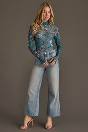 A woman with long blonde hair stands against a gray background, smiling slightly as she wears the Panache Apparel Collection’s Teal, Cream & Light Pink Floral Mesh Top paired with light blue high-waisted wide-leg jeans and nude heeled sandals.