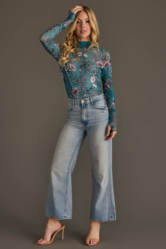 A woman models the Panache Apparel Collection Teal, Cream & Light Pink Floral Mesh Top, paired with high-waisted light blue wide-leg jeans and nude heeled sandals. She poses against a plain gray background, with one hand touching her hair.