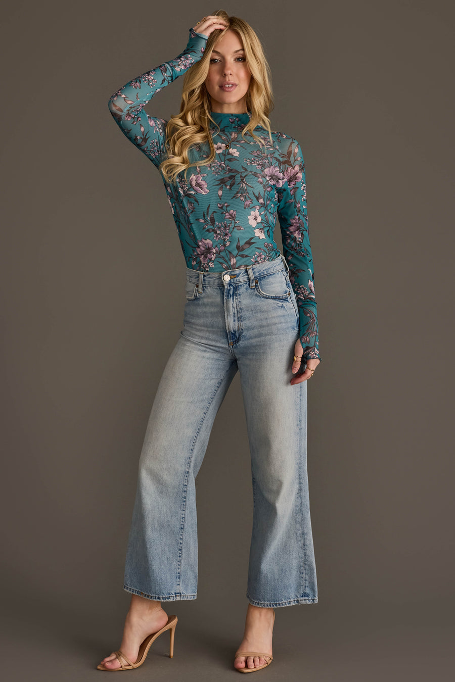 A woman models the Panache Apparel Collection Teal, Cream & Light Pink Floral Mesh Top, paired with high-waisted light blue wide-leg jeans and nude heeled sandals. She poses against a plain gray background, with one hand touching her hair.