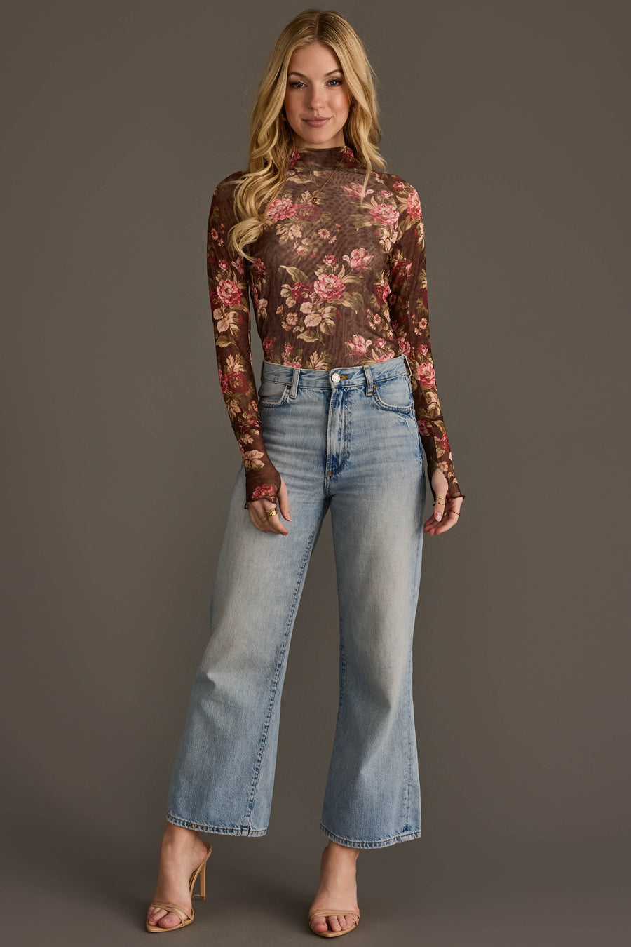A woman stands against a plain background, wearing the Panache Apparel Collection Brown, Cream & Burgundy Floral Mesh Top paired with high-waisted, light blue wide-leg jeans. She has long blonde hair and is styled with heeled sandals.