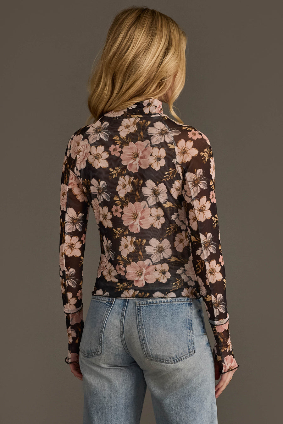 A woman with long blonde hair, seen from behind, wears the Panache Apparel Collection’s Black & Light Pink Floral Mesh Top paired with light blue high-waisted jeans against a plain background.
