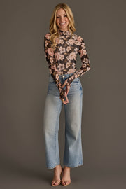 A woman stands against a plain background, smiling. She wears the Panache Apparel Collection’s Black & Light Pink Floral Mesh Top with long sleeves, paired with high-waisted light blue jeans and open-toed heels. Her long hair is loose and wavy.