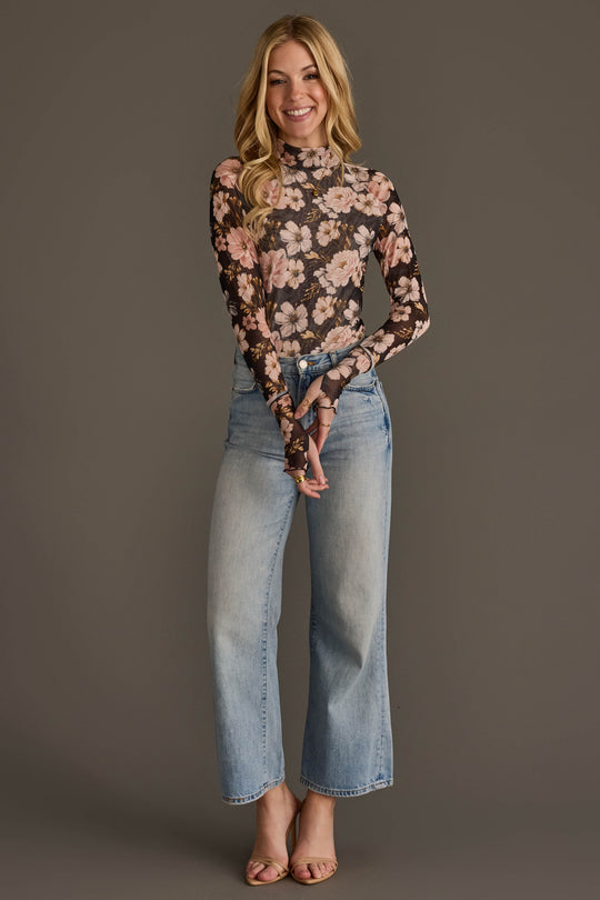 A woman stands against a plain background, smiling. She wears the Panache Apparel Collection’s Black & Light Pink Floral Mesh Top with long sleeves, paired with high-waisted light blue jeans and open-toed heels. Her long hair is loose and wavy.