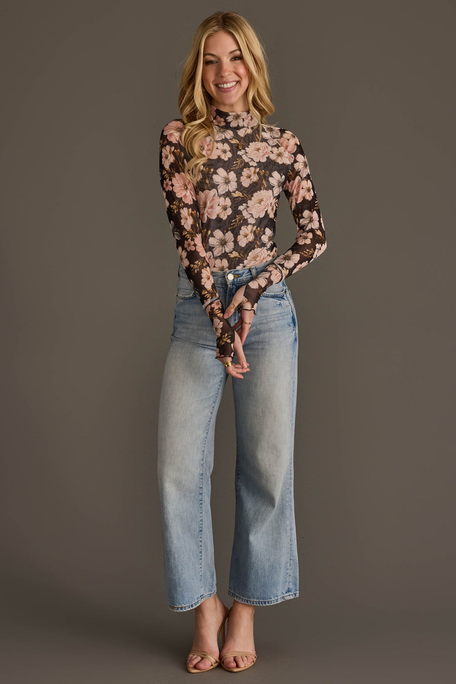 A woman stands against a plain background, smiling. She wears the Panache Apparel Collection’s Black & Light Pink Floral Mesh Top with long sleeves, paired with high-waisted light blue jeans and open-toed heels. Her long hair is loose and wavy.