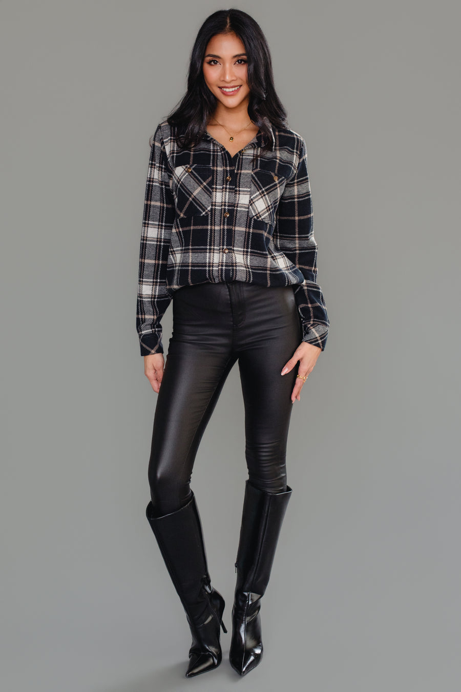 A woman stands smiling, wearing the Panache Apparel Collection Black, White & Tan Plaid Button Up, paired with black leather pants and tall black boots against a plain gray background.