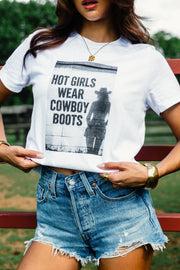 A woman stands outdoors by a red fence, wearing the Panache Apparel Collection’s “Hot Girls Wear Cowboy Boots Graphic Tee” featuring a cowgirl image and bold text, paired with denim shorts.