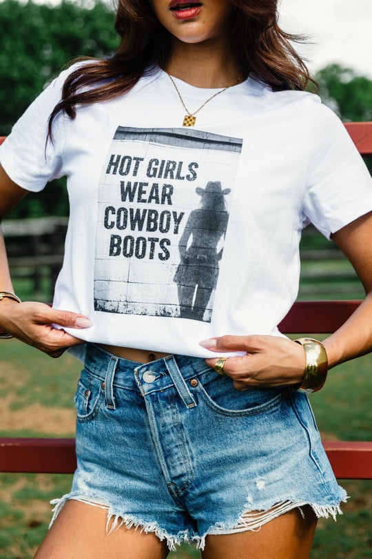 A woman stands outdoors by a red fence, wearing the Panache Apparel Collection’s “Hot Girls Wear Cowboy Boots Graphic Tee” featuring a cowgirl image and bold text, paired with denim shorts.