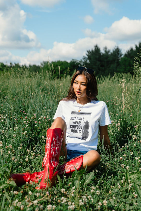 A woman relaxes in a grassy field beneath a blue, cloud-dotted sky, wearing the Panache Apparel Collection’s Hot Girls Wear Cowboy Boots Graphic Tee paired with denim shorts and bold red cowboy boots. Trees provide a picturesque backdrop to the scene.