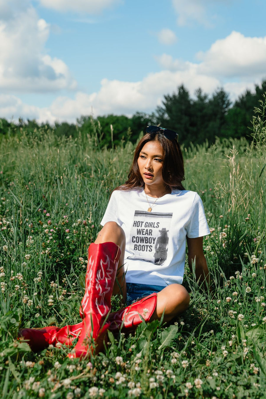 A woman relaxes in a grassy field beneath a blue, cloud-dotted sky, wearing the Panache Apparel Collection’s Hot Girls Wear Cowboy Boots Graphic Tee paired with denim shorts and bold red cowboy boots. Trees provide a picturesque backdrop to the scene.