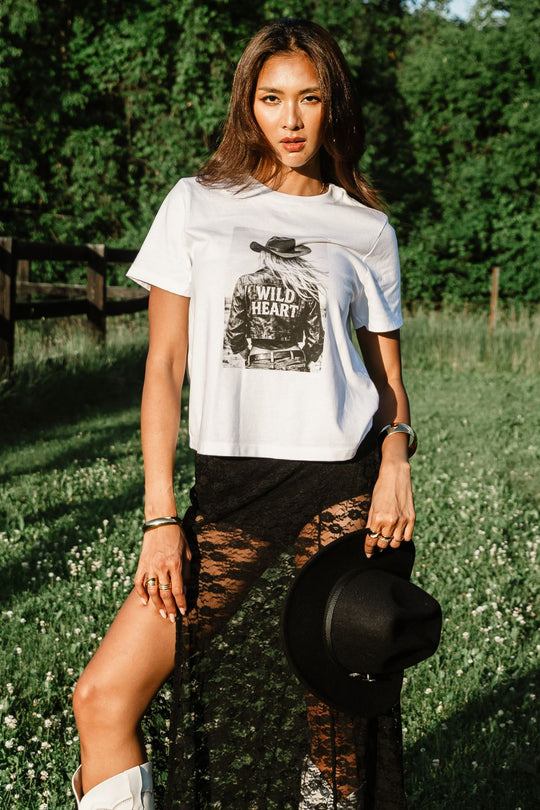 A woman stands outdoors in the sunlight, wearing the Panache Apparel Collection’s Wild Heart Graphic Tee—crafted from soft Airlume cotton—paired with a sheer black skirt and holding a black hat in one hand. Lush green trees and grass form the background.