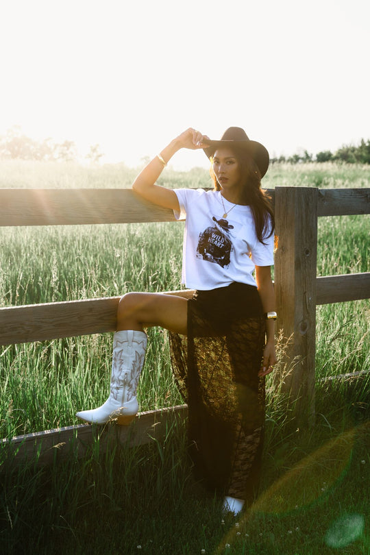 A woman in a cowboy hat, Panache Apparel Collection’s Wild Heart Graphic Tee crafted from soft Airlume cotton, a sheer black skirt, and white boots leans on a wooden fence in a sunny grassy field.