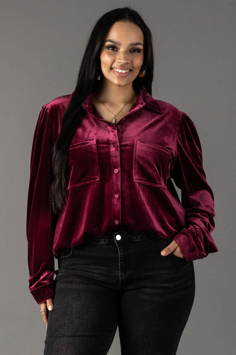 A smiling woman wears a long-sleeved maroon velvet button-up shirt from the Panache Apparel Collection featuring two chest pockets, paired with black jeans.