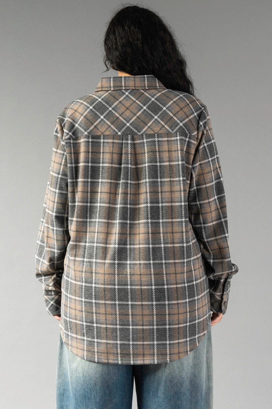 A person with long dark hair, dressed in the Panache Apparel Collection Gray, Tan & White Plaid Button Up and blue jeans, is standing with their back to the camera against a plain gray background.