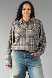 A woman with long curly hair wears the Gray, Tan & White Plaid Button Up from Panache Apparel Collection with blue jeans, standing against a plain gray background.