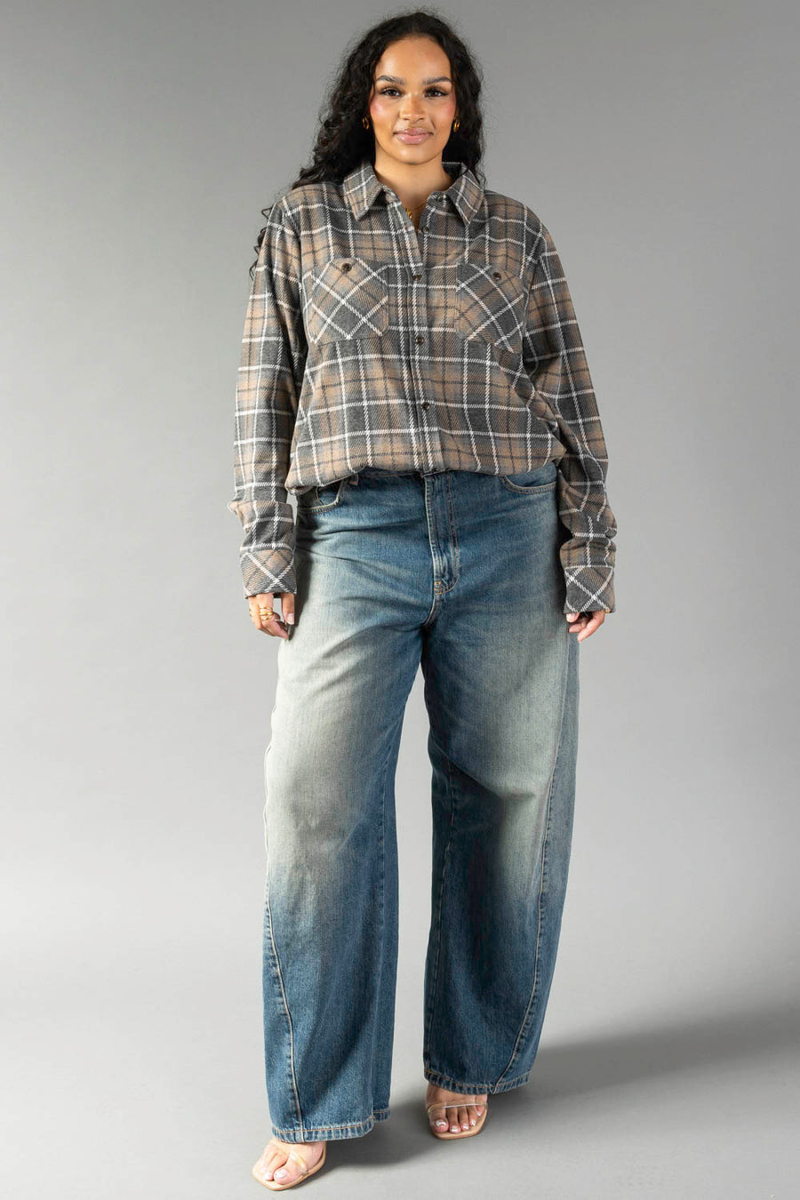 A woman stands against a gray background wearing the Panache Apparel Collection’s Gray, Tan & White Plaid Button Up. She pairs the shirt with loose-fitting faded blue jeans and open-toe sandals. Her long, wavy hair frames her face as she smiles slightly.