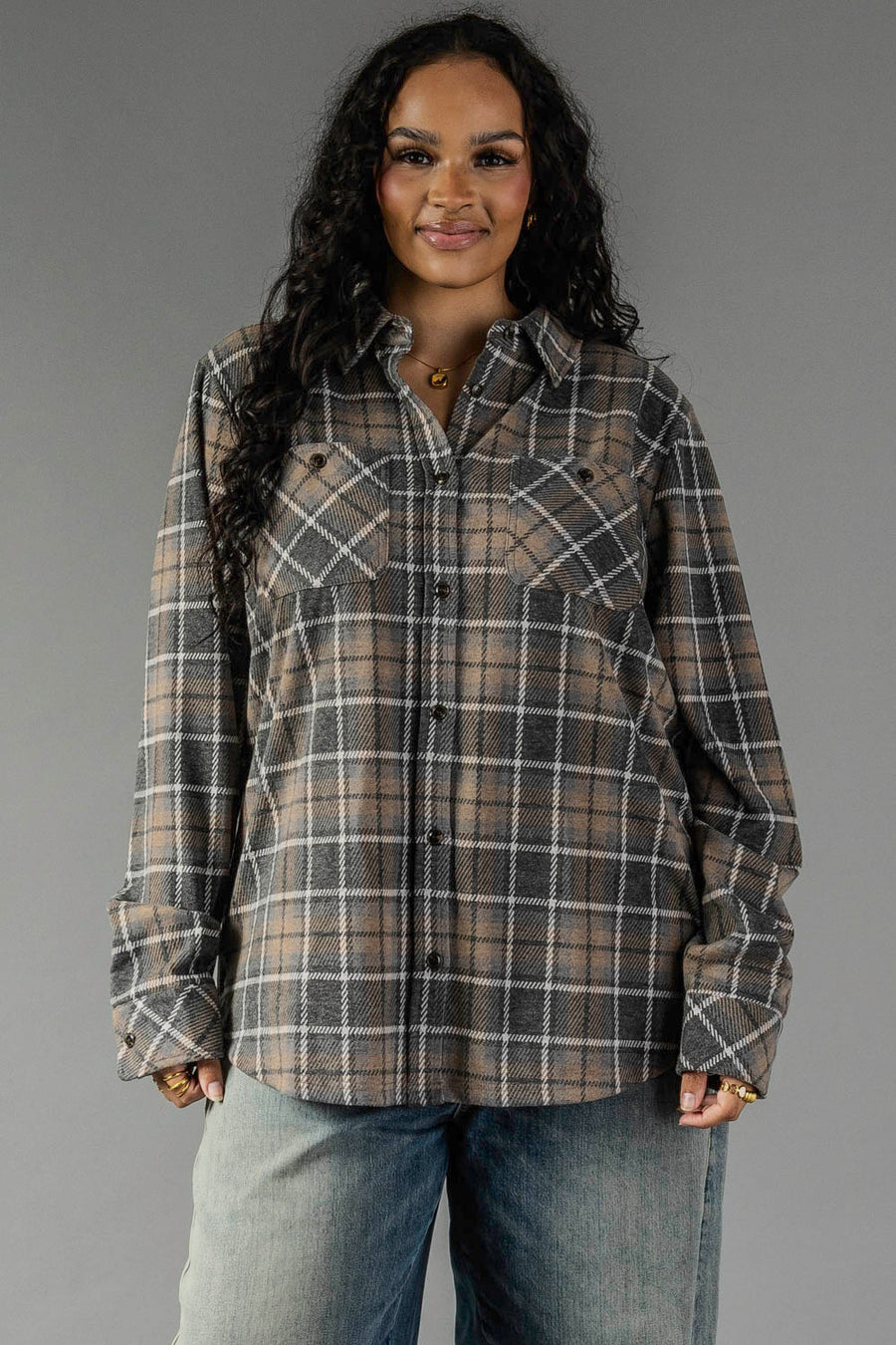 A woman with long curly hair wears the Panache Apparel Collection Gray, Tan & White Plaid Button Up and light blue jeans, standing against a plain gray background and smiling slightly.
