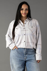 A woman with long dark hair wears the Panache Apparel Collection’s White, Gray & Light Blue Plaid Button Up, featuring two chest pockets, paired with blue jeans. She stands with one hand in her pocket against a plain gray background.