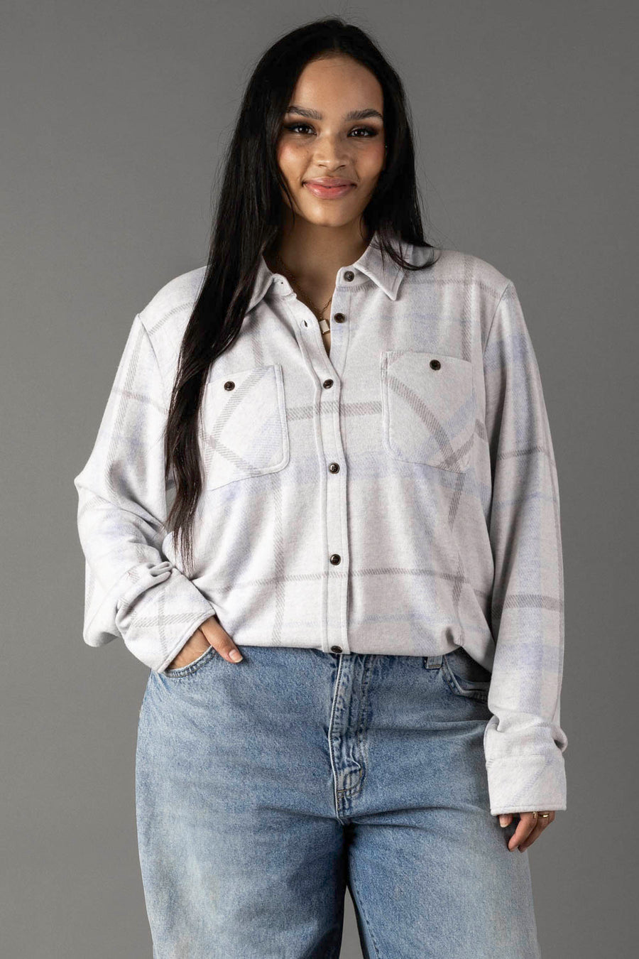 A woman with long dark hair wears the Panache Apparel Collection’s White, Gray & Light Blue Plaid Button Up, featuring two chest pockets, paired with blue jeans. She stands with one hand in her pocket against a plain gray background.