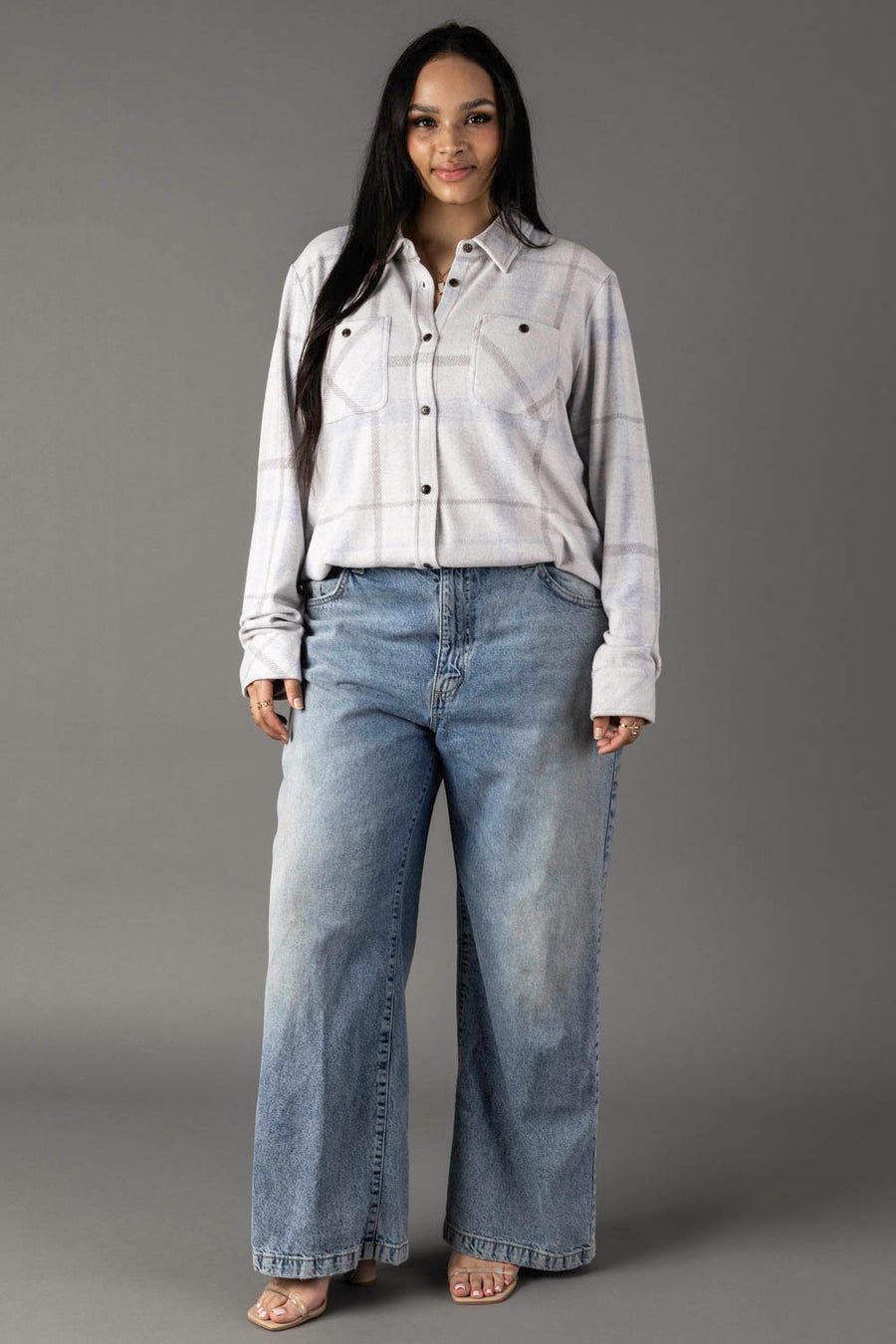 A woman with long dark hair poses in a relaxed manner against a gray background, wearing the Panache Apparel Collection’s White, Gray & Light Blue Plaid Button Up paired with wide-leg blue jeans and open-toe shoes.