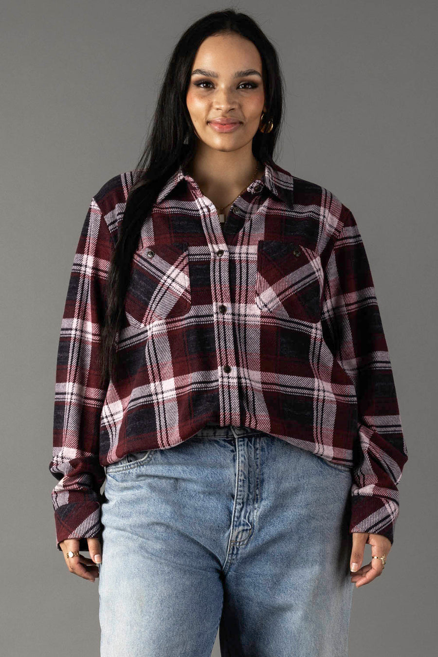 A woman with long dark hair, dressed in the Panache Apparel Collection’s Maroon, Charcoal & White Plaid Button Up and light blue jeans, stands against a plain gray background, smiling slightly at the camera.