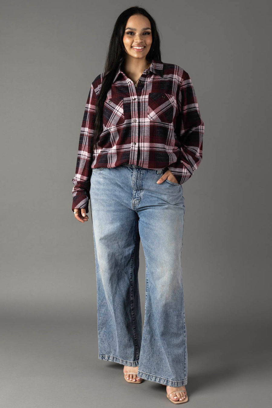 A person stands smiling against a plain gray background, wearing the Panache Apparel Collection’s Maroon, Charcoal & White Plaid Button Up, wide-leg blue jeans, and sandals, with one hand in their pocket.