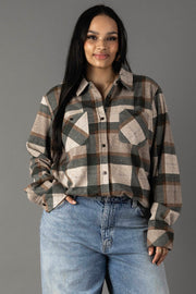 A woman with long dark hair wears the Panache Apparel Collection’s Tan, Dark Green & Rust Plaid Button Up, paired with light blue jeans. She stands against a plain gray background, smiling softly at the camera.