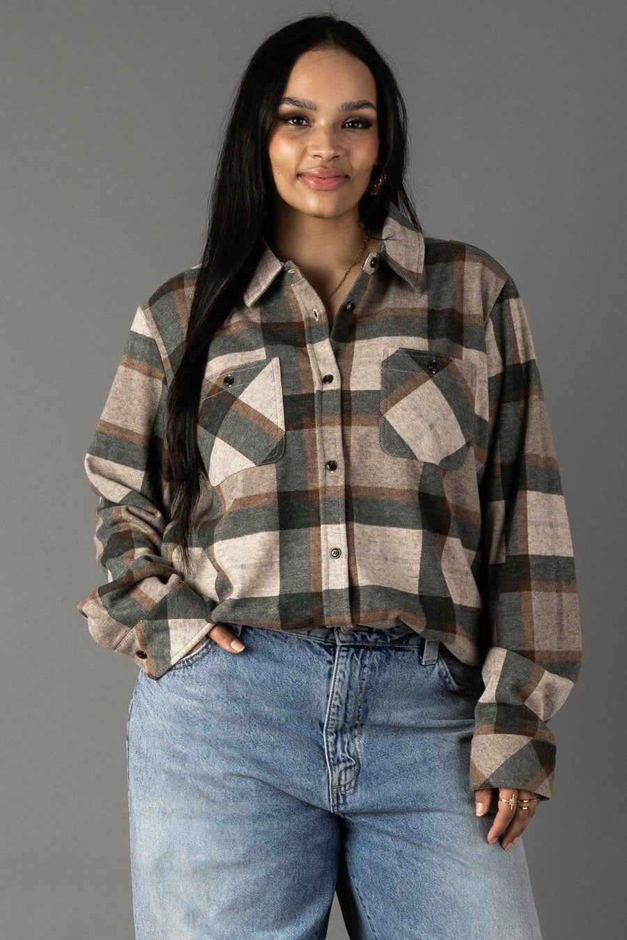 A woman with long dark hair wears the Panache Apparel Collection’s Tan, Dark Green & Rust Plaid Button Up, paired with light blue jeans. She stands against a plain gray background, smiling softly at the camera.