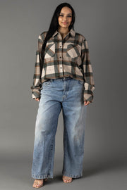 A woman stands against a gray background, wearing the Panache Apparel Collection’s Tan, Dark Green & Rust Plaid Button Up and loose-fitting blue jeans. She has long dark hair and is smiling slightly, with her hands relaxed at her sides.
