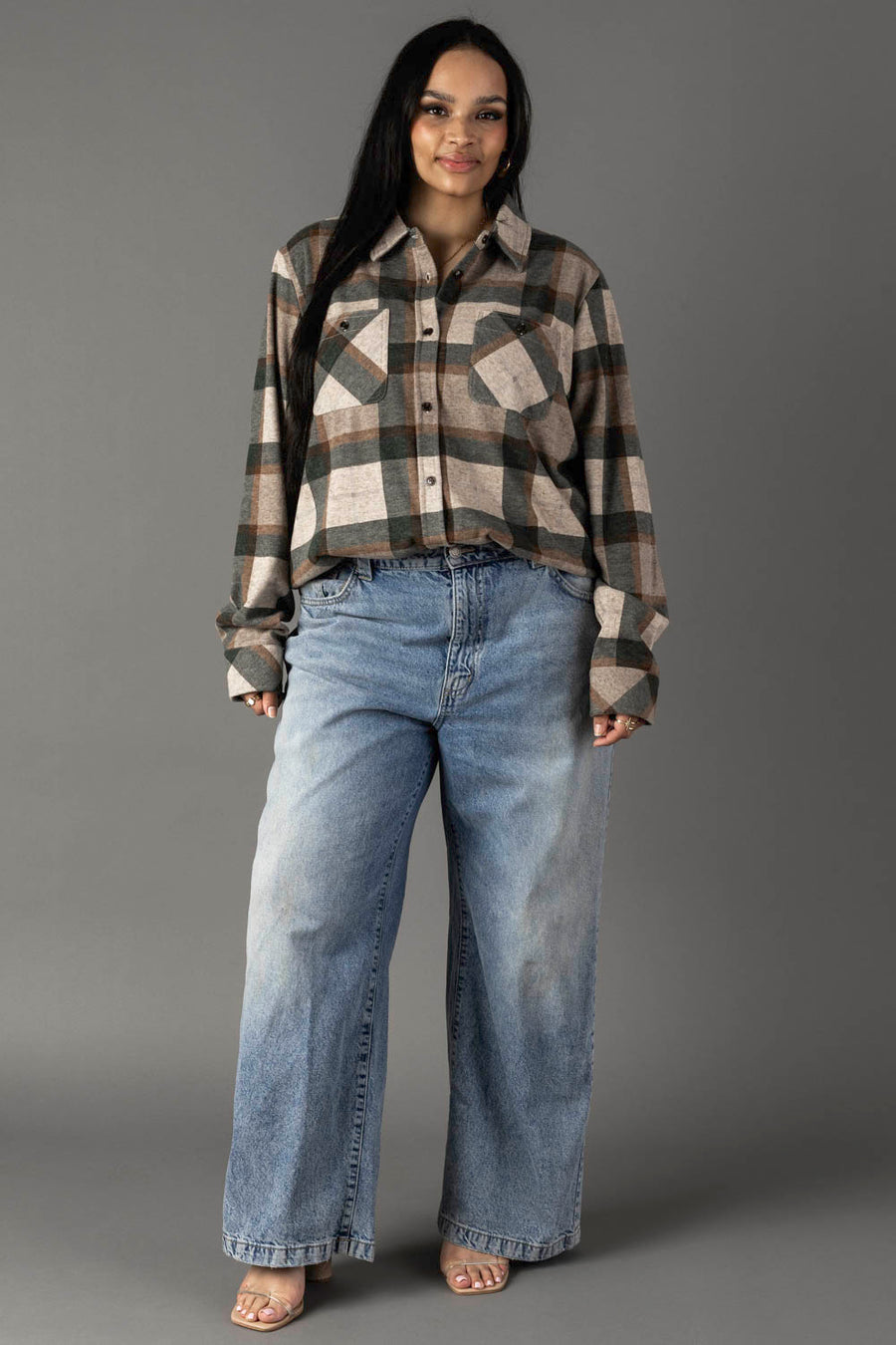 A woman stands against a gray background, wearing the Panache Apparel Collection’s Tan, Dark Green & Rust Plaid Button Up and loose-fitting blue jeans. She has long dark hair and is smiling slightly, with her hands relaxed at her sides.
