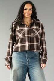 A woman with long curly hair wears the Panache Apparel Collection’s Dark Brown, Cream & Black Plaid Button Up and blue jeans, standing and smiling in front of a plain gray background.