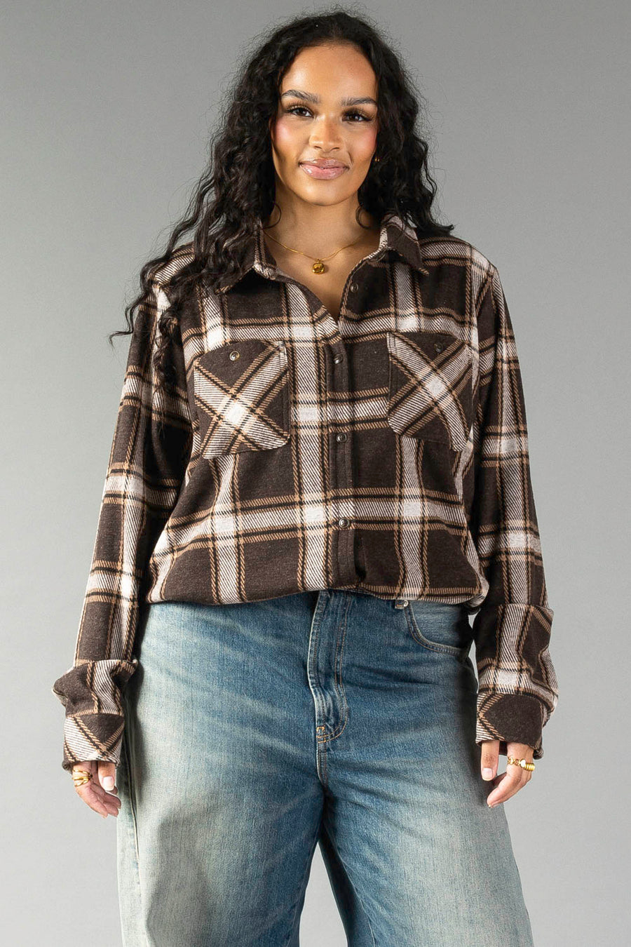 A woman with long curly hair wears the Panache Apparel Collection’s Dark Brown, Cream & Black Plaid Button Up and blue jeans, standing and smiling in front of a plain gray background.