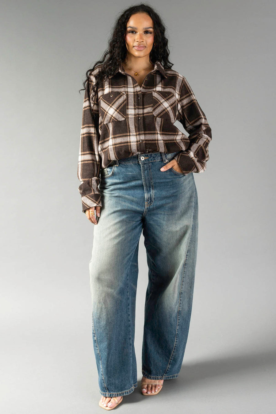A person with long dark hair wears the Dark Brown, Cream & Black Plaid Button Up from Panache Apparel Collection and wide-leg blue jeans, standing against a plain gray background.